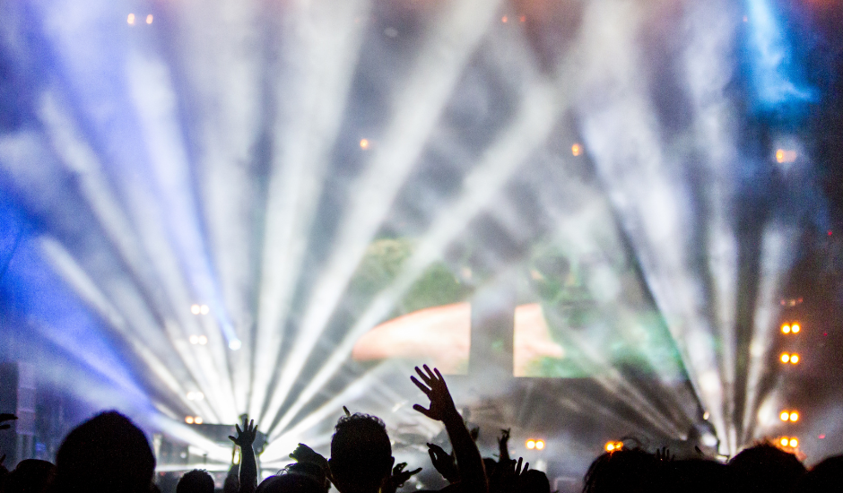 lights on a stage shine whilst the audience is in the dark, arms in the air